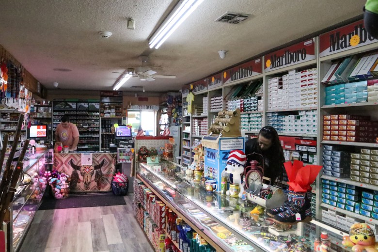 The Osage Trading Co. viewed from the inside. Two employees stand behind counters in a narrow space, tobacco products along one wall and other items for sale.