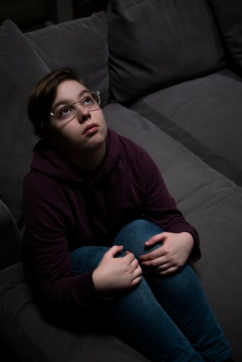 Hannah Norris sits on a dark gray sofa with her legs bent and her hands on her knees. She is wearing jeans, a purple hoodie and glasses. She has short brown hair.
