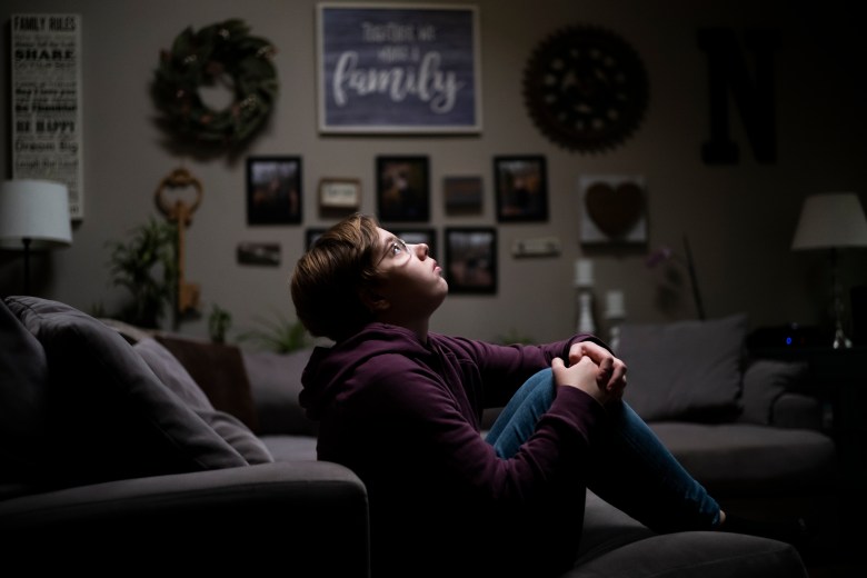 Hannah Norris sits on a dark gray sofa with her legs bent and her hands on her knees. She is wearing jeans, a purple hoodie and glasses. She has short brown hair.
