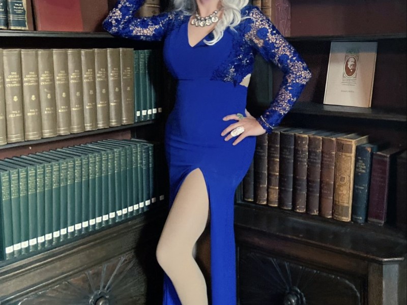Lady J wears a long blue dress with lace sleeves and a white necklace. She stands in front of two bookcases filled with books. She has long blond hair and black and yellow eye makeup with dark lipstick."