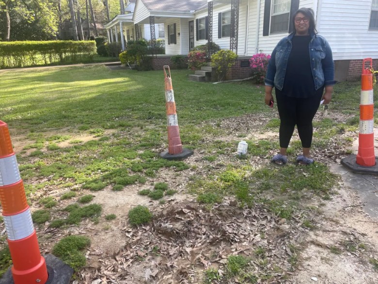 Amia Edwards is standing on her lawn outside her home beside orange-and-white cones marking the repairs she needed from flooding sewage.