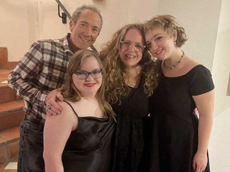 The author, Amy Silverman poses with her family at the base of a staircase. In the front is Sophie who has long hear, wears glasses and is wearing a sleeveless black dress. Amy's husband Ray Stern is behind Sophie. He wears a plaid shirt and jeans. He has his right hand on Sophie's right shoulder. Amy is in the middle. She has long wavy hair and wears glasses. She also is wearing a black dress. On the right, is Annabelle, who has her arm around Amy. She has short hair and is wearing a black dress.