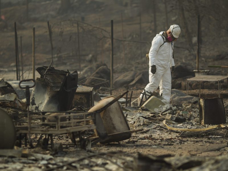 Someone walk through the rubble of a fire in a forest. Burned tables and chairs are seen thrown about in the scene. Trees are burned, too.
