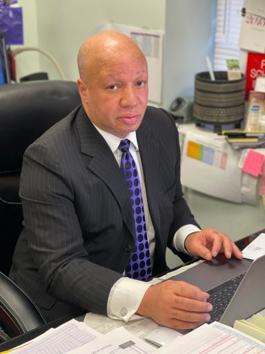 De Lacy Davis is a bald man. He's sitting at a desk with a laptop and papers in front of him. He wears a gray suit, a white shirt and a blue tie.