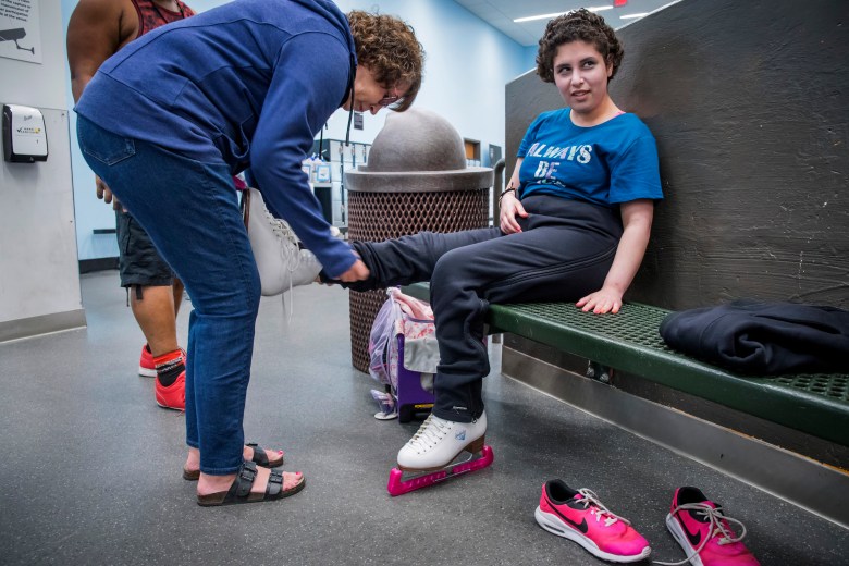 Zainab Edwards sits on a green bench while her mom ties her right ice skate. She is wearing black pants and a blue t-shirt. Her mom is wearing jeans and a blue sweatshirt.   