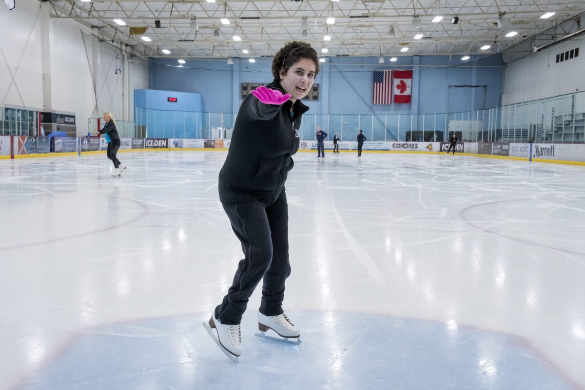 Zainab Edwards points her right arm forward while standing on the ice. She is wearing black pants, a black sweatshirt and pink gloves.