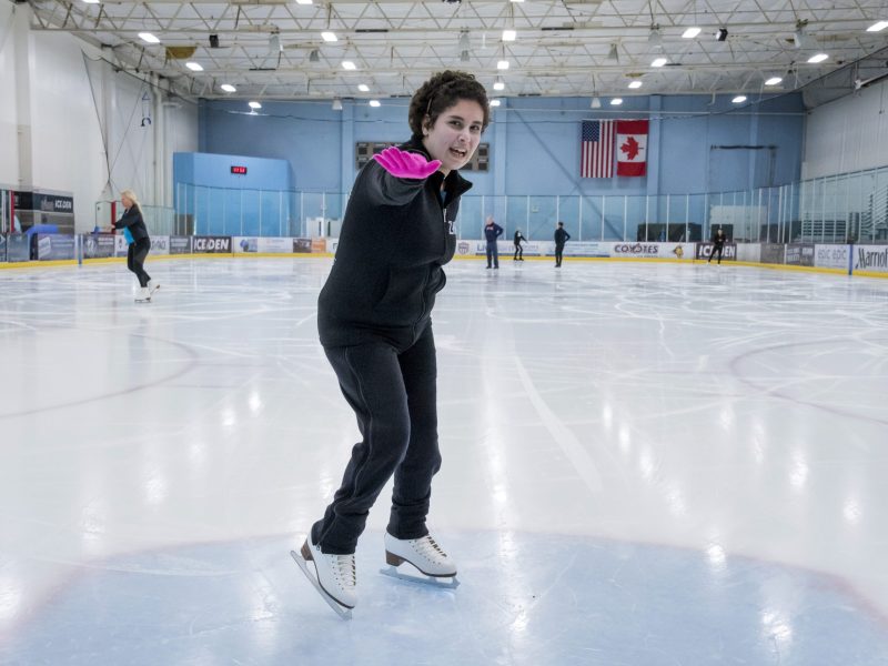 Zainab Edwards points her right arm forward while standing on the ice. She is wearing black pants, a black sweatshirt and pink gloves.