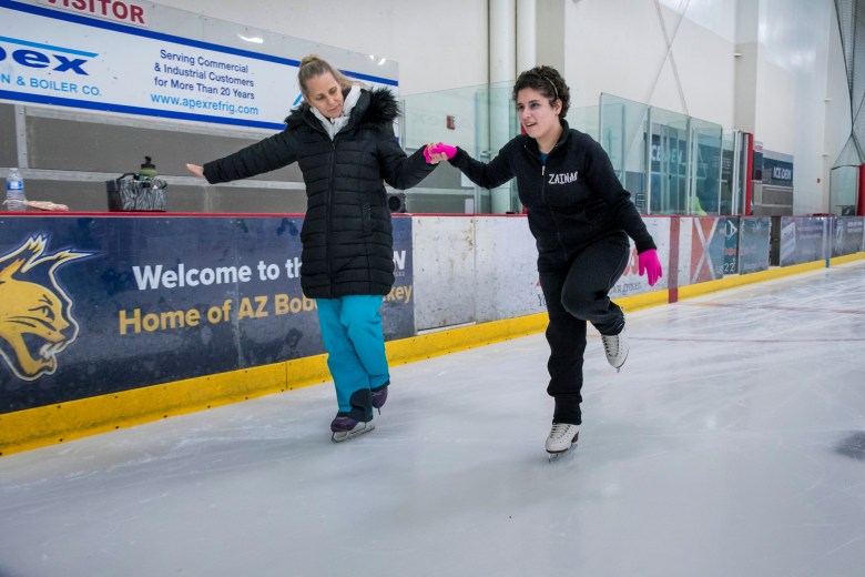 Zainab Edwards holds hands with her teacher Tammy Jimenez as they skate. Zainab wears a black sweatshirt with her name on the upper left. Her left knee is bent. Her instructor wears blue pants and a black coat. She is watching Zainab as they skate.