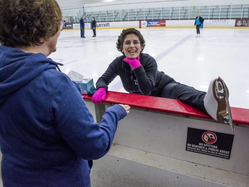 Zainab Edwards puts her left leg on the edge of the ice rink to stretch as her mom talks with her. Zainab has her hand under her chin and is smiling.