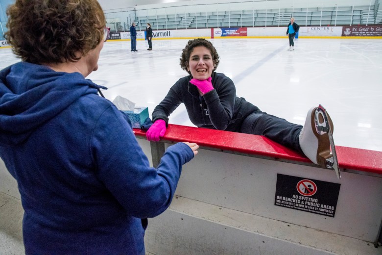 Zainab Edwards stretches during a skating lesson. She wears black pants, a black sweatshirt and pink gloves. Her left foot is on rail of the rink. Her mom is talking with her. Zainab's mom is wearing a blue sweatshirt.