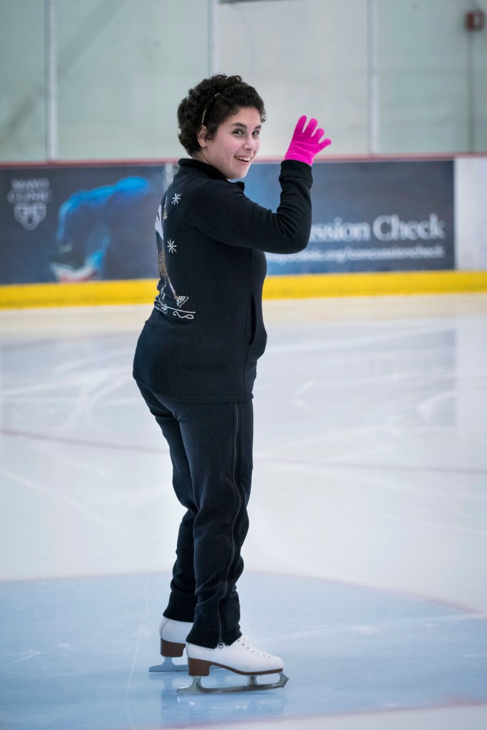 Zainab Edwards waves while standing on the ice. She is wearing black pants, a black sweatshirt and pink gloves.  She has short dark hair and is smiling.