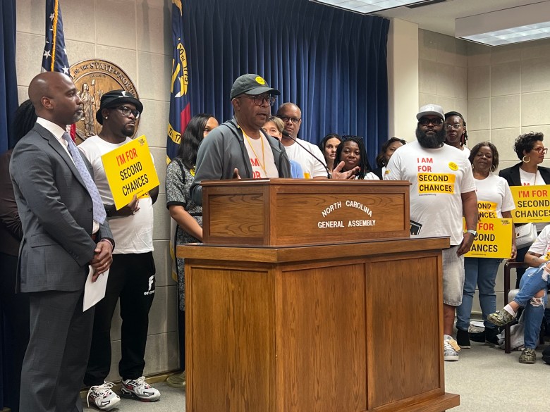 Dennis Gaddy stands in front of a podium with several people standing around him with signs that say "I'm ready for second chances."