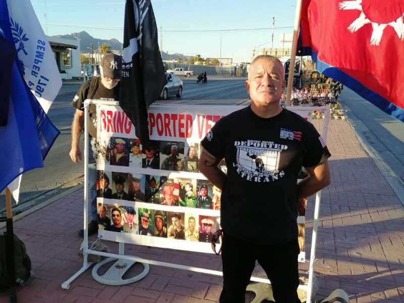 Marcelino Ramos, a 54 year old man, stands in front of a banner that has flags behind it and photos of deported veterans on the front. He is wearing a black tshirt and black pants.