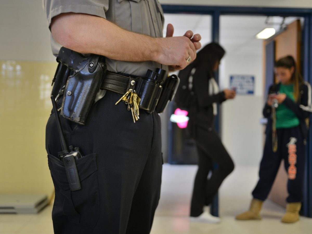 a police officer stands in a school hallway. Two students stand behind him out of focus. The viewer can see the items on his belt like keys, mace, and his holster.