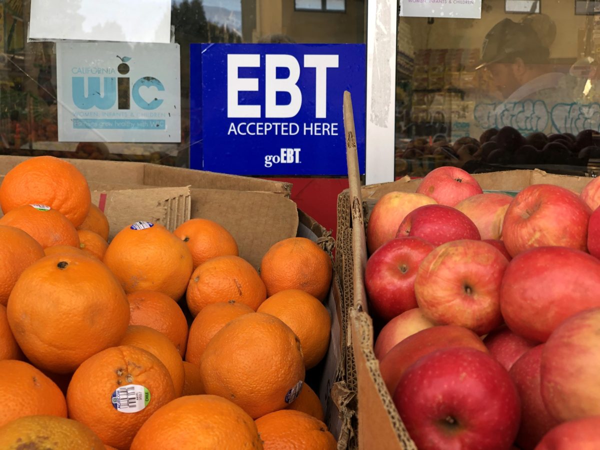 A box of oranges and a box of apples are displayed outside of a grocery store. On the window is a sign that says the store accepts EBT and WIC.