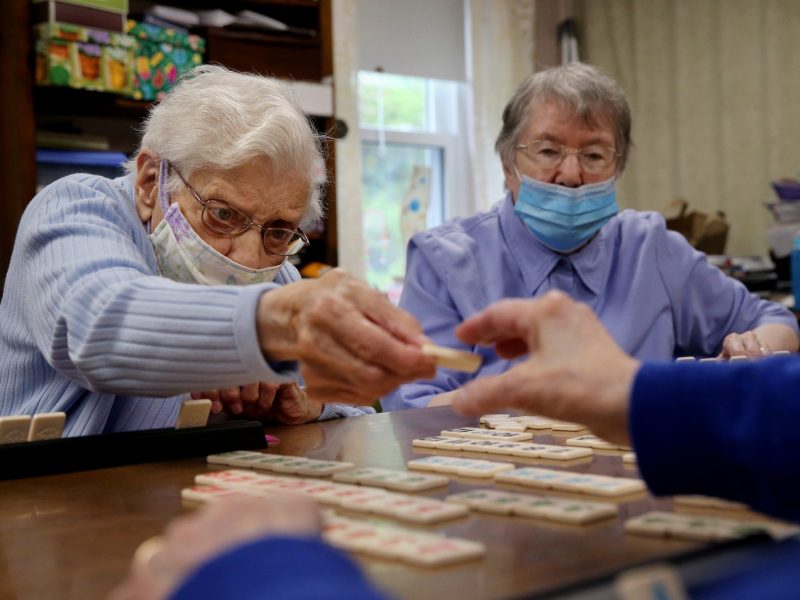 two nursing home residents play a board game at a table. they both wear blue paper face masks.