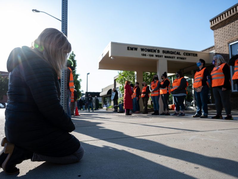 A pro-life demonstrator kneels before a line of volunteer clinic escorts.