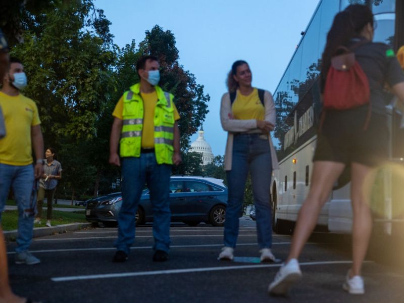 Two men and two women stand outside a bus in D.C. with the Capitol dome in the background. The two men look at the bus door. They wear surgical masks. They wear yellow t-shirts and jeans, one wears a safety vest. Another woman looks on with her arms folded. She wears a yellow t-shirt, a white sweater and jeans. Another woman faces away,looking in the bus door. She wears dark shorts and a dark t-shirt. She wears a red backpack.