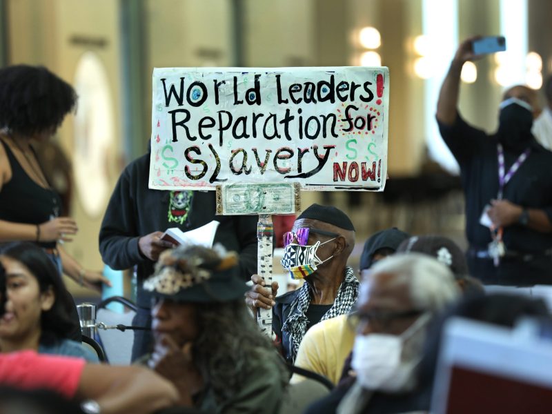 A seated man wearing a mask holds a sign that says "World leaders! Reparations for slavery now!" during a California reparations task force meeting.