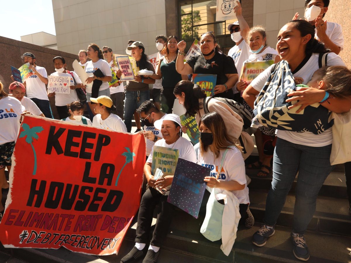 A crowd of people, all wearing white tshirts and holding a red sign saying "Keep LA Housed" stand together in front of a building.
