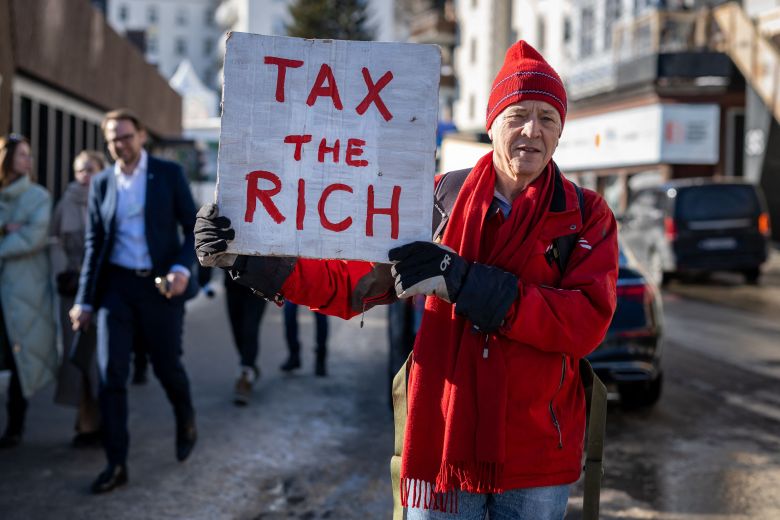 Phil White is holding a hand-lettered sign on white cardboard that reads, "Tax the Rich." He's wearing a red hat and coat.