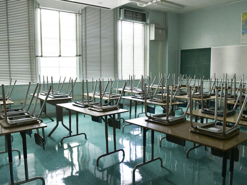 In an empty classroom, seats are upside down on desks as the sun shines in the windows.