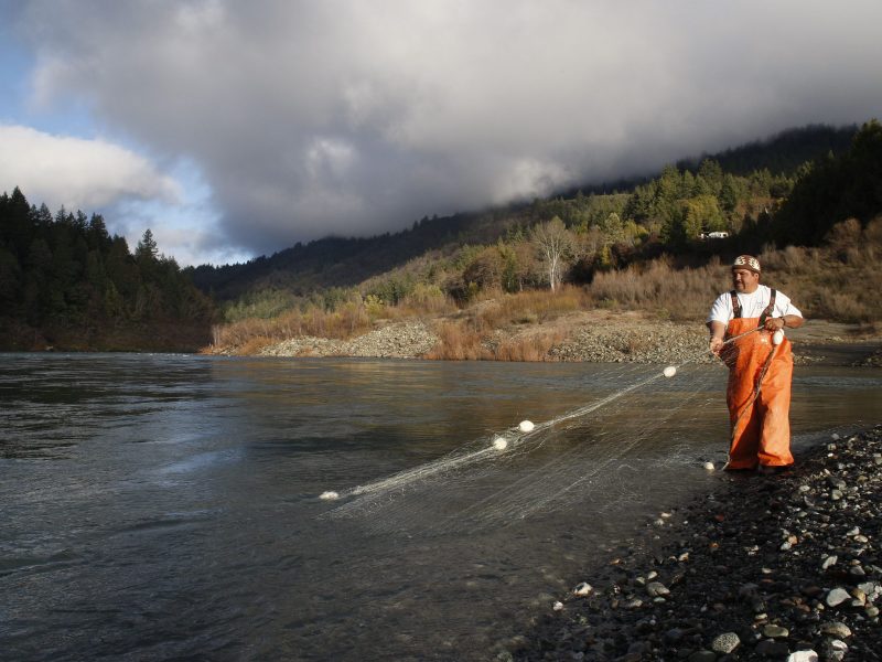 A Yurok tribe member wearing orange coveralls checks the net they have placed in a large body of water. there are small hills behind them and a cloudy sky.