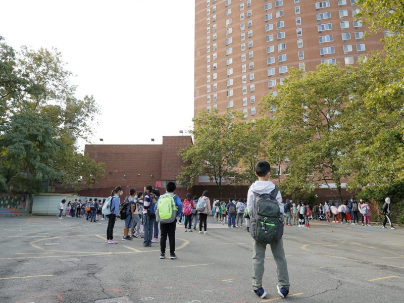 Students stand and walk along the blacktop toward red brick school buildings. There are green trees that line the way.