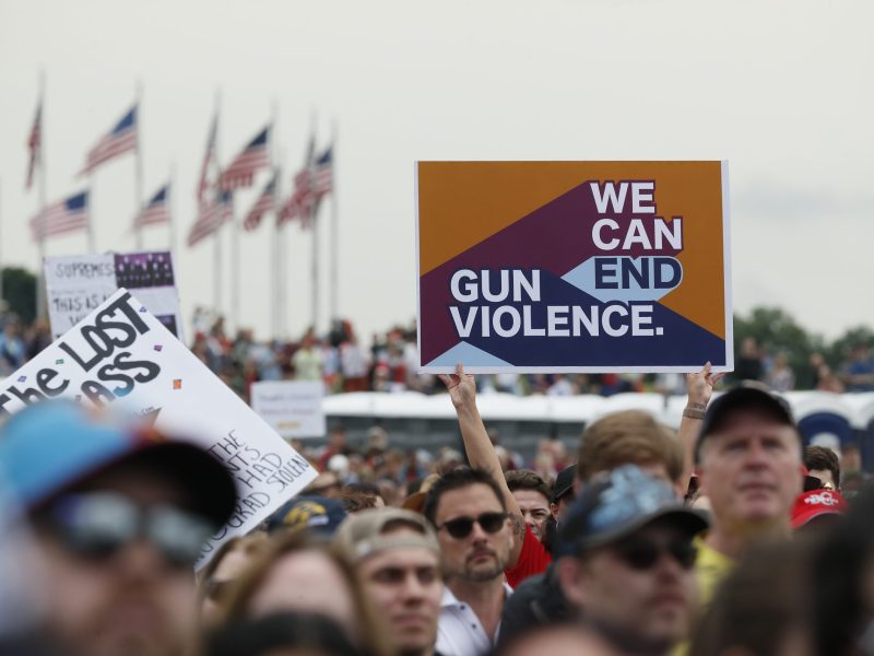 A crowd of people at a gun violence protest, their faces are out of focus. One sign that reads 'We Can End Gun Violence' is held up above the crowd.