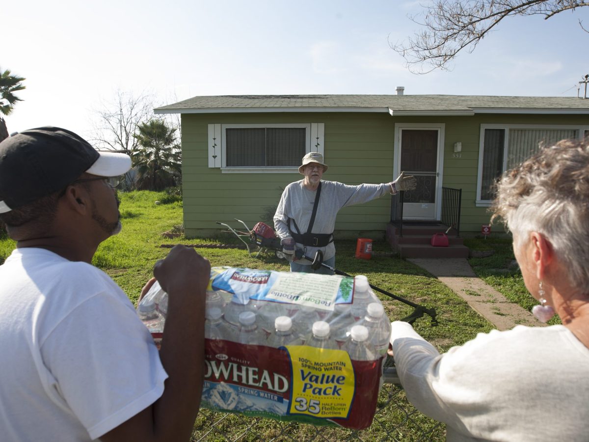 The three people identified in the caption are standing outside a small, green ranch-style house, two holding a pack of water bottles.