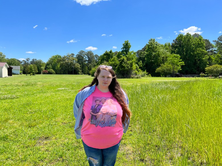 Heather French wears jeans, a blue shirt and a pink T-shirt with the cast of the Golden Girls and the words "Stay Golden" written on it. She stands with her hands behind her a tree-lined field. 