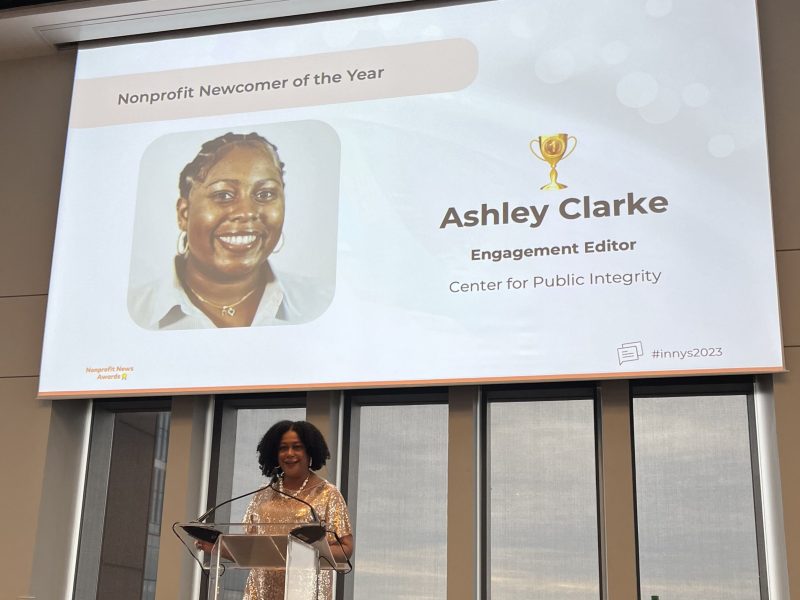 A woman stands at a podium below the picture of another woman on a screen that says "Nonprofit Newcomer of the Year: Ashley Clarke, Engagement Editor, Center for Public Integrity."