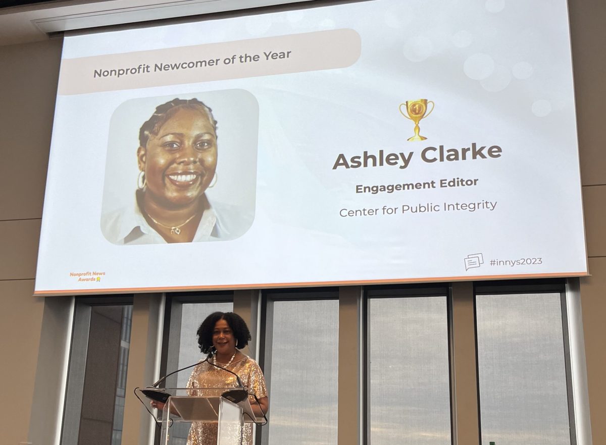 A woman stands at a podium below the picture of another woman on a screen that says "Nonprofit Newcomer of the Year: Ashley Clarke, Engagement Editor, Center for Public Integrity."