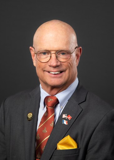Steven Holt wears glasses and is dressed in a dark blazer with several pins fastened to his blazer. 