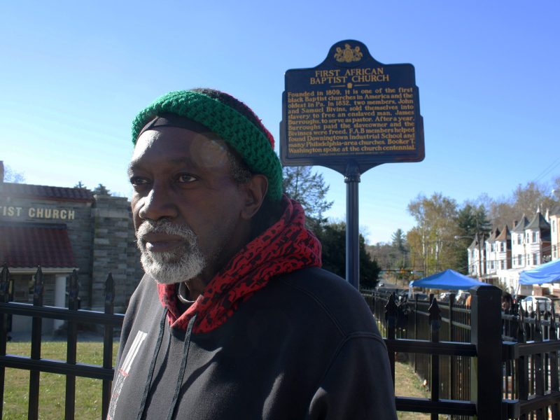 A man (Jondhi Harrell) stands in front of a sign for a church. He is wearing a hat, scarf, and coat.
