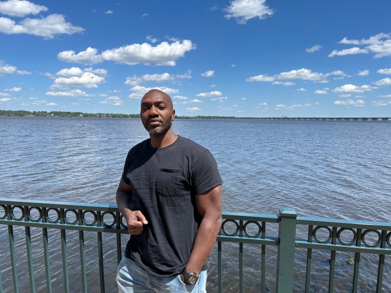 Kevin Slade poses by a body of water. He is wearing a black t-shirt and jeans. 