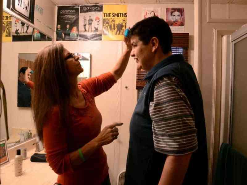 Mark smiles at his mother as she brushes his hair. They're in the bathroom, posters in the background.