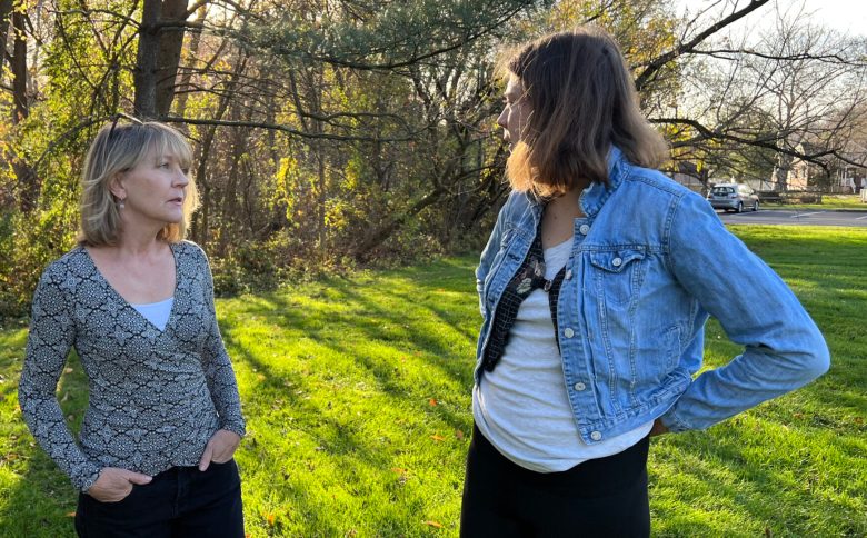 Two women talking. Woman on right has dark shoulder-length hair. She's wearing a denim jacket and white shirt. The other woman has blond shoulder-length hair and is wearing a blue sweater. They are outside on green grass with trees in the background.