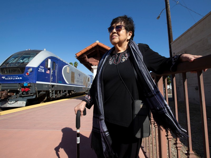 Mary Acosta Rodriguez Martinez Garcia is wearing sunglasses and looking up, leaning on metal fencing and her cane, beside railroad tracks with a Pacific Surfliner train on them.