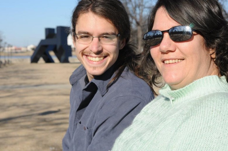 Joshua Atkins, on the left, smiles with his mother, Lauren, on the right. Joshua is wearing a blue shirt and glasses and Lauren is wearing sunglasses and a green sweater.