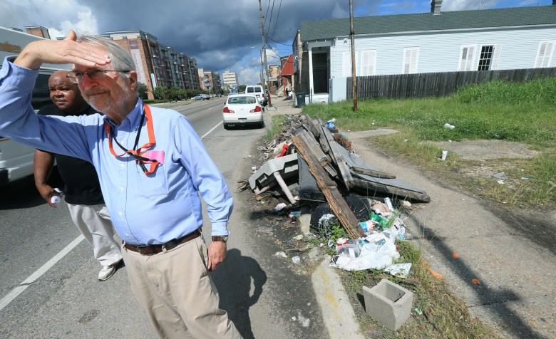Professor Howard Mielke of Tulane University stands in the street as he looks into the distance with his hand over his eyes so he can block the sun. Trash and debris lie on the curb beside him.