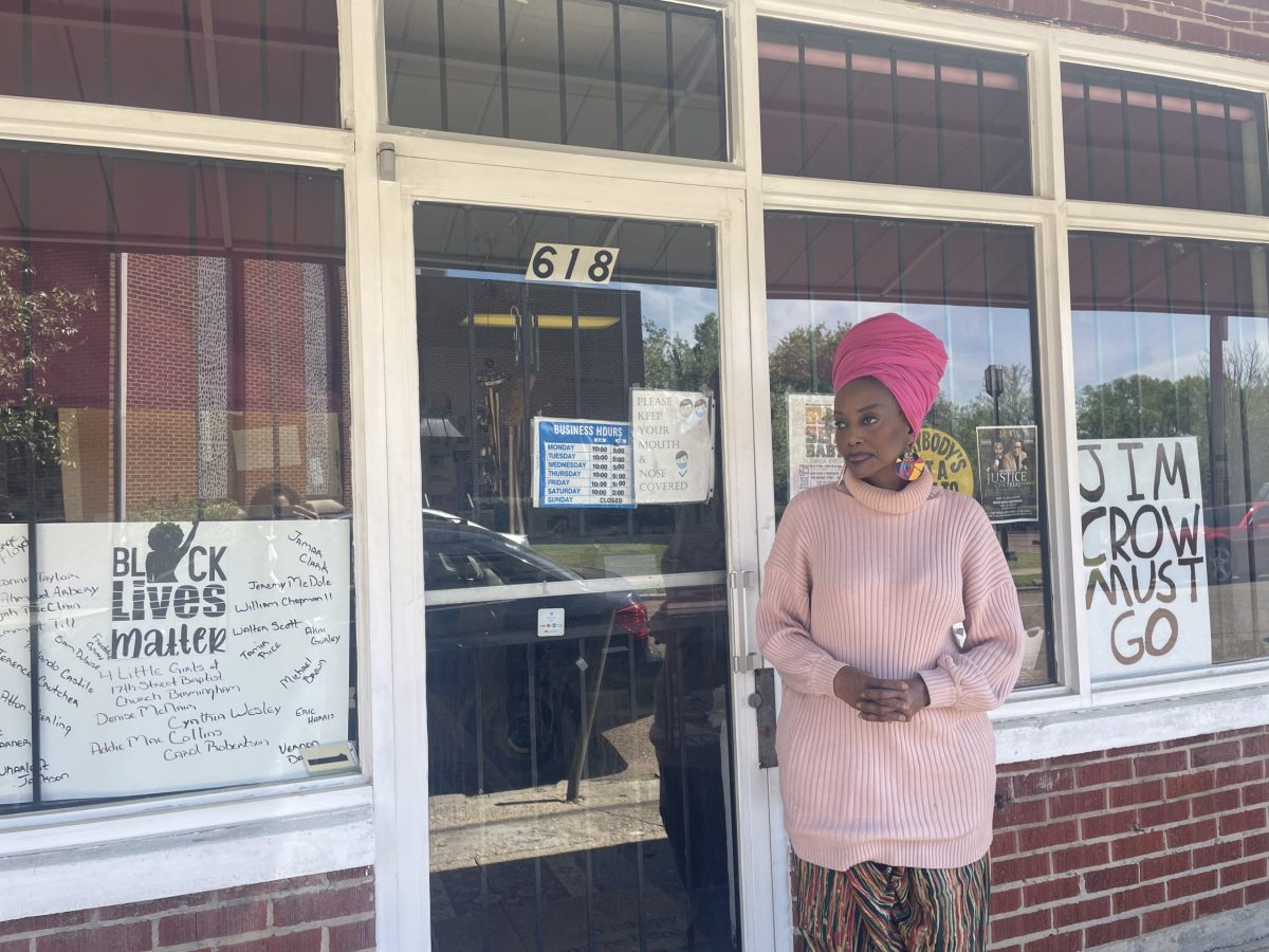 Maati Jone Primm stands in front of her store. She is wearing a pink outfit, and she has two signs in the windows of her store. One says "Jim Crow Must Go" and the other says "Black Lives Matter."