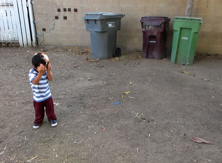 Ruben, 2, plays hide and seek in his front yard. He is surrounded by dirt on the ground and trash bins. 