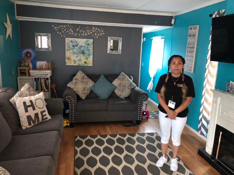 Patty Flores stands in the living room of her mobile home. She wears a black shirt and white pants and her hair is in two braids. The walls are painted blue.