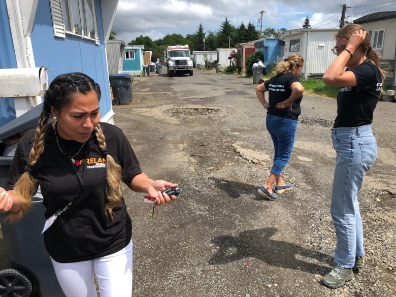 Patty Flores wears a black shirt and white pants as she stands in front of her blue mobile home with white trimming. Her two colleagues are standing next to her, with their backs facing the cameras. There are potholes in the road.