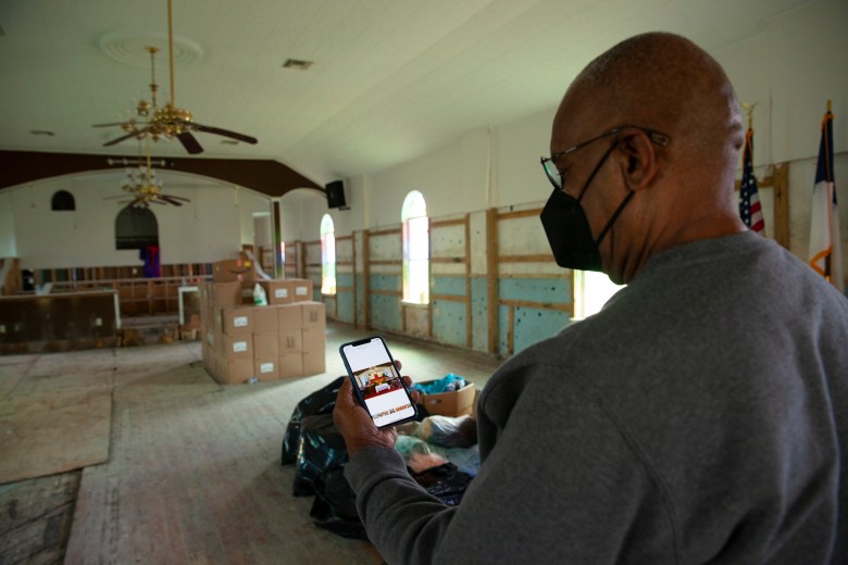 The Rev. Haywood Johnson Jr. has a mask around his face as he stands inside the damaged Saint Paul Missionary Baptist Church while he looks at his phone which has an older photo of the church before it was damaged.