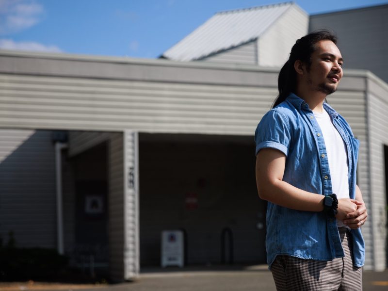 MJ Dizon stands outside the North Thurston Public Schools' Family & Youth Resource Center in a blue button down shirt. Dizon has long black hair and is smiling.